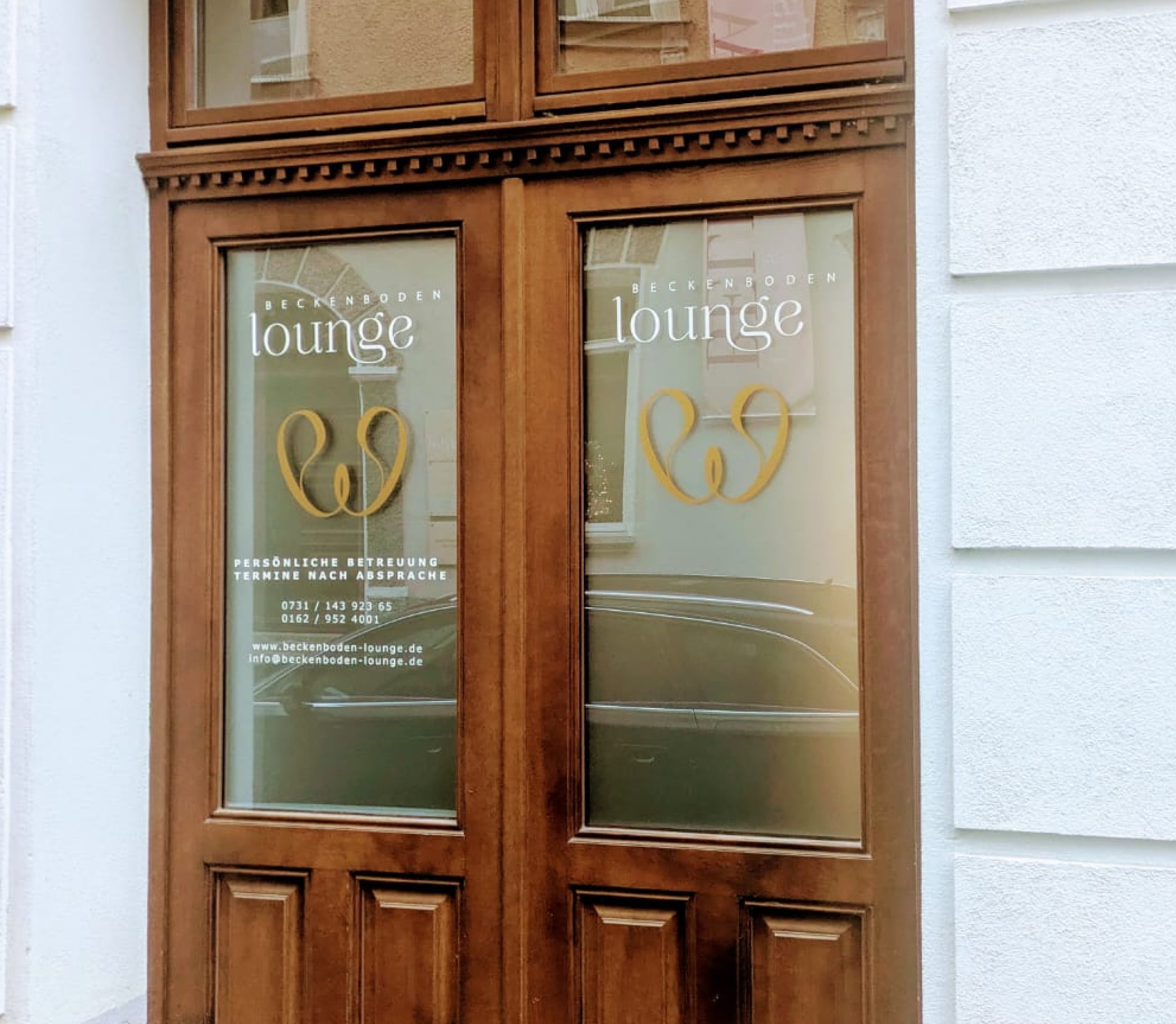 Eingang der Beckenboden Lounge mit eleganter Holztür und beschrifteten Glasfenstern.